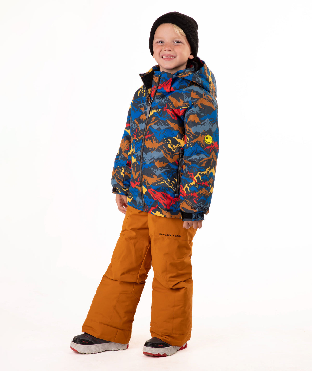 A child wearing a colorful mountain patterned hooded jacket, rust-orange snow pants, a black beanie, and sneakers, standing against a plain white studio background.