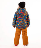 A child wearing a colorful mountain patterned hooded jacket, rust-orange snow pants, a black beanie, and sneakers, standing against a plain white studio background.