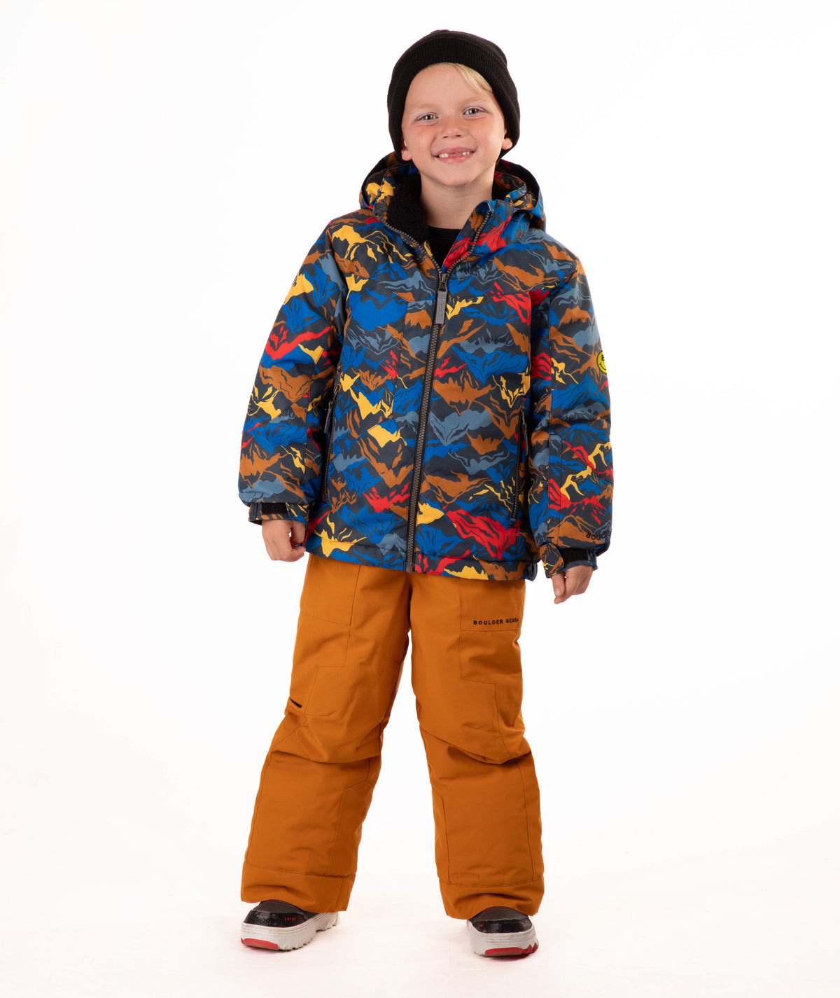 A child wearing a colorful mountain patterned hooded jacket, rust-orange snow pants, a black beanie, and sneakers, standing against a plain white studio background.