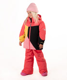 A young girl wearing a color-blocked ski jacket in pink, coral, yellow, and black, matching snow pants, a pink beanie, standing against a white studio background.