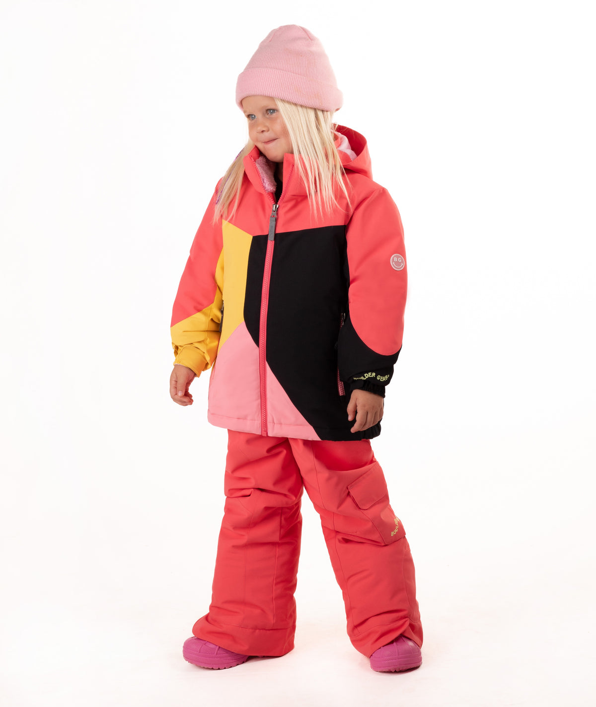 A young girl wearing a color-blocked ski jacket in pink, coral, yellow, and black, matching snow pants, a pink beanie, standing against a white studio background.