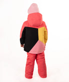A young girl wearing a color-blocked ski jacket in pink, coral, yellow, and black, matching snow pants, a pink beanie, standing against a white studio background.