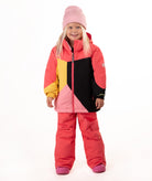 A young girl wearing a color-blocked ski jacket in pink, coral, yellow, and black, matching snow pants, a pink beanie, standing against a white studio background.