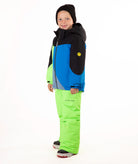 A child wearing a color-blocked ski jacket and matching snow pants, standing against a plain white studio background. The jacket has black, blue, and neon green panels, and the pants are bright neon green.