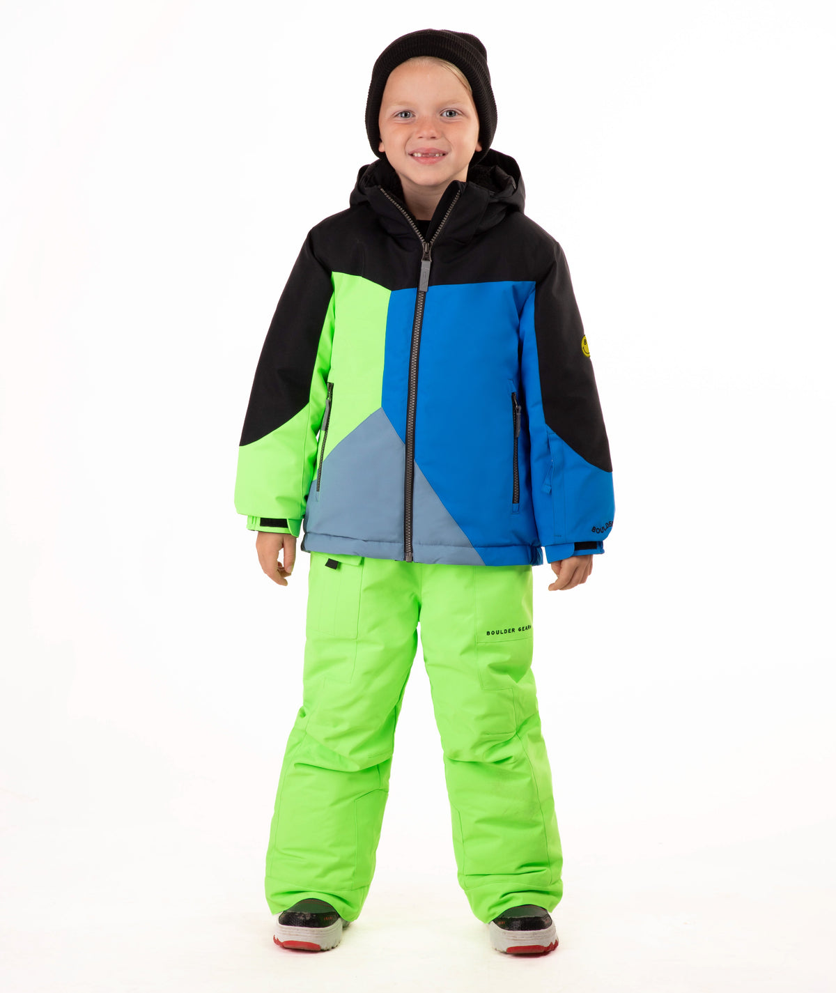 A child wearing a color-blocked ski jacket and matching snow pants, standing against a plain white studio background. The jacket has black, blue, and neon green panels, and the pants are bright neon green.