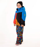 A young child wearing a color-blocked ski jacket in blue, red, black, and orange, paired with patterned snow pants and a black beanie, standing against a white studio background.