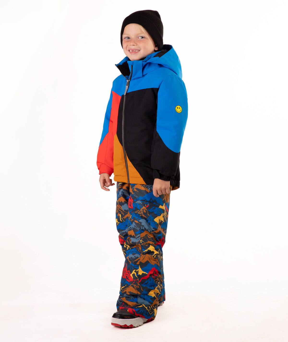 A young child wearing a color-blocked ski jacket in blue, red, black, and orange, paired with patterned snow pants and a black beanie, standing against a white studio background.