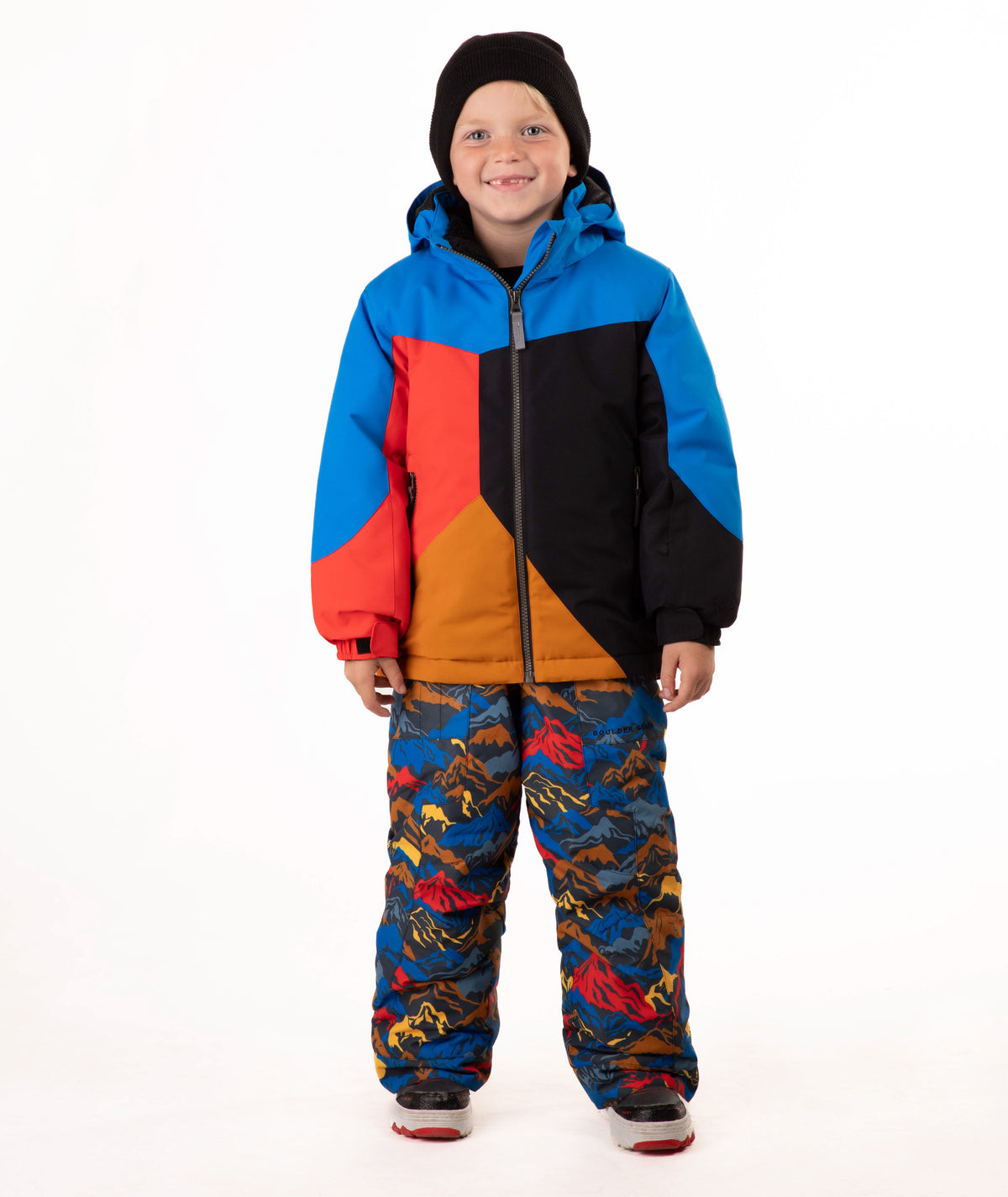 A young child wearing a color-blocked ski jacket in blue, red, black, and orange, paired with patterned snow pants and a black beanie, standing against a white studio background.