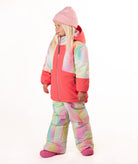 A young girl wearing a pink tie-dye jacket, consisting of a bright pink jacket and matching pants, along with a pink beanie and pink boots, standing against a white studio background.