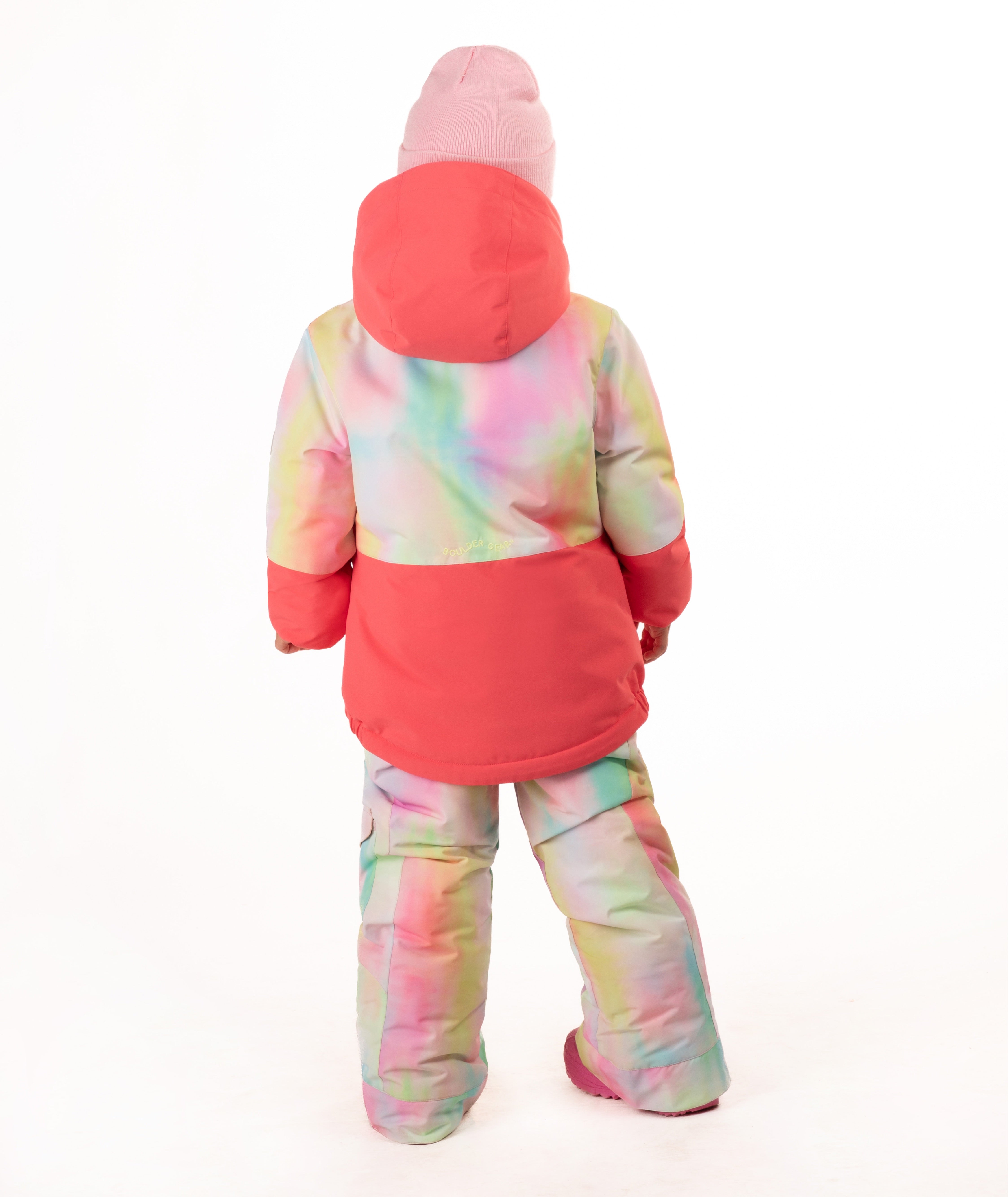 A young girl wearing a pink tie-dye jacket, consisting of a bright pink jacket and matching pants, along with a pink beanie and pink boots, standing against a white studio background.