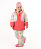 A young girl wearing a pink tie-dye jacket, consisting of a bright pink jacket and matching pants, along with a pink beanie and pink boots, standing against a white studio background.