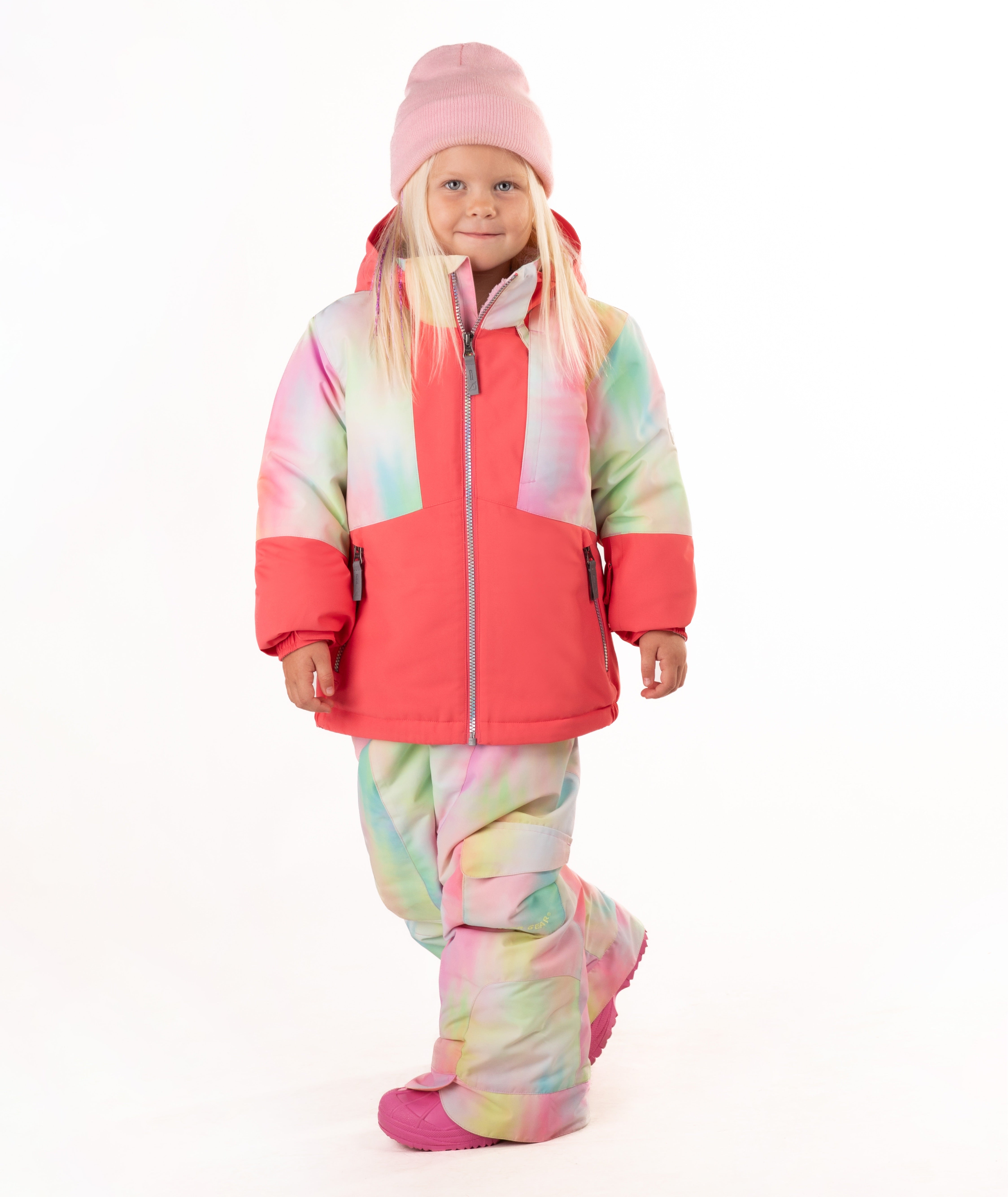 A young girl wearing a pink tie-dye jacket, consisting of a bright pink jacket and matching pants, along with a pink beanie and pink boots, standing against a white studio background.