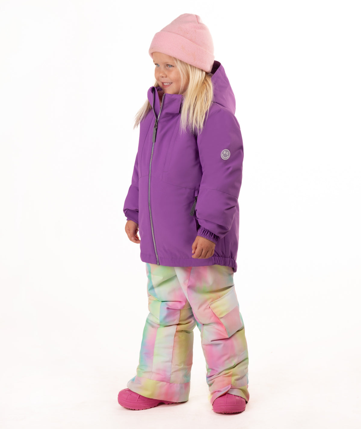 A young girl smiling and posing in a purple winter jacket, pastel tie-dye snow pants, a pink beanie, and pink boots against a solid white background.