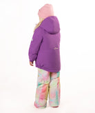A young girl smiling and posing in a purple winter jacket, pastel tie-dye snow pants, a pink beanie, and pink boots against a solid white background.