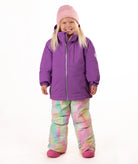 A young girl smiling and posing in a purple winter jacket, pastel tie-dye snow pants, a pink beanie, and pink boots against a solid white background.
