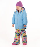 A young child wearing a light blue winter jacket, a purple beanie, and colorful geometric print snow pants, standing against a plain white background.