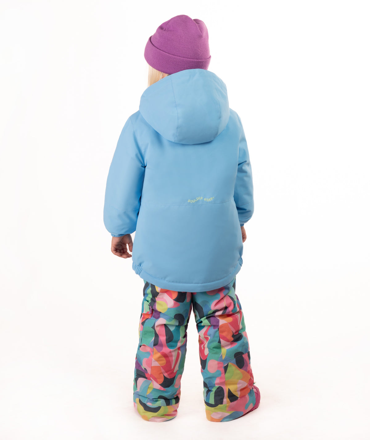 A young child wearing a light blue winter jacket, a purple beanie, and colorful geometric print snow pants, standing against a plain white background.
