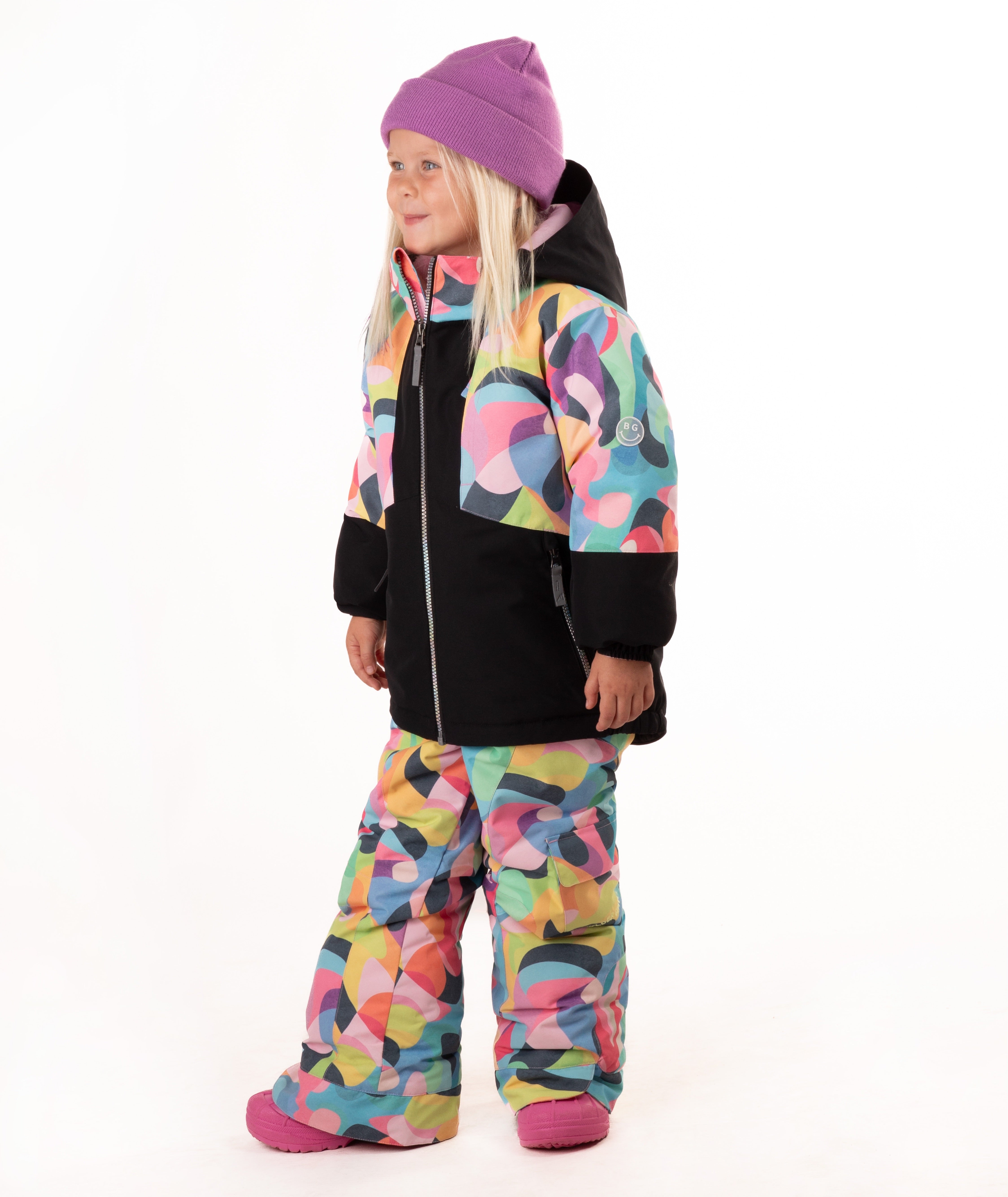 A young girl wearing a colorful abstract print ski jacket and matching pants, a purple beanie, and pink boots, standing against a white studio background.