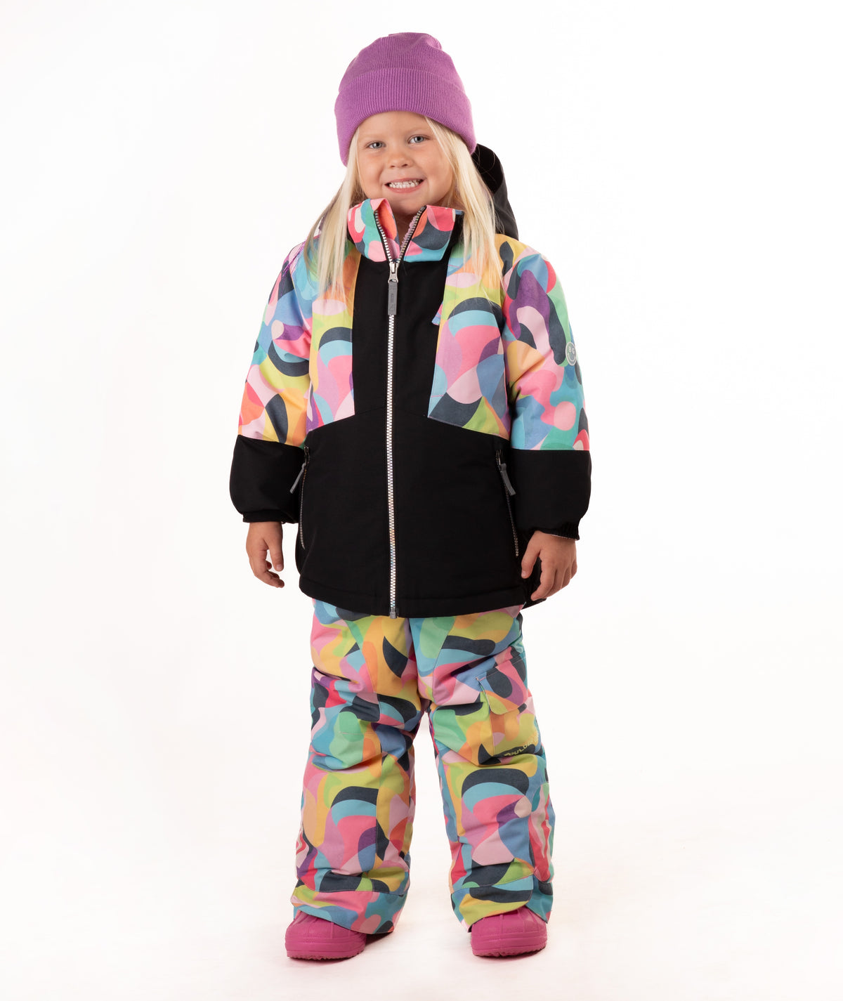 A young girl wearing a colorful abstract print ski jacket and matching pants, a purple beanie, and pink boots, standing against a white studio background.