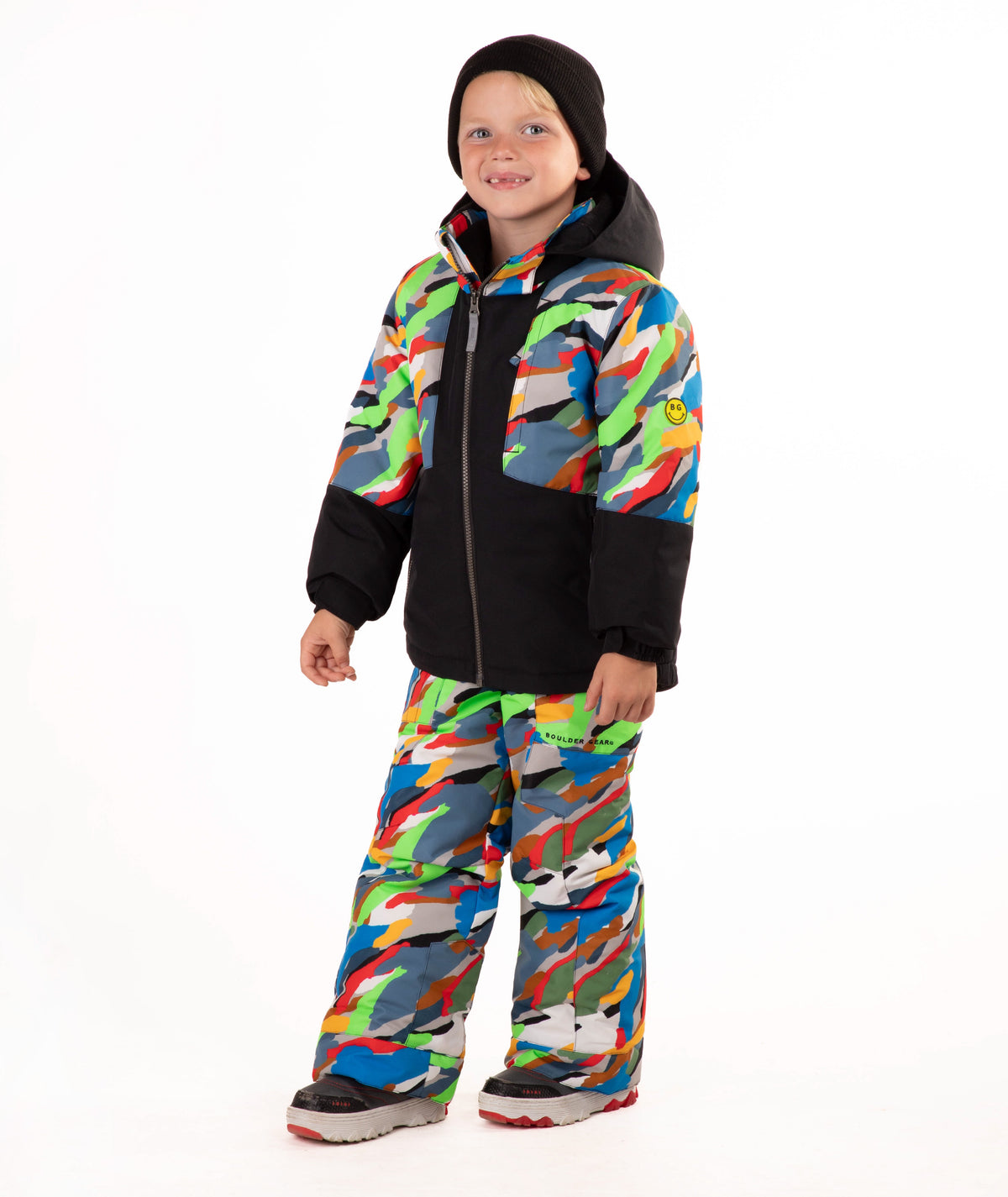 A child wearing a colorful camouflage ski jacket and matching pants with a black beanie, standing against a plain white background.