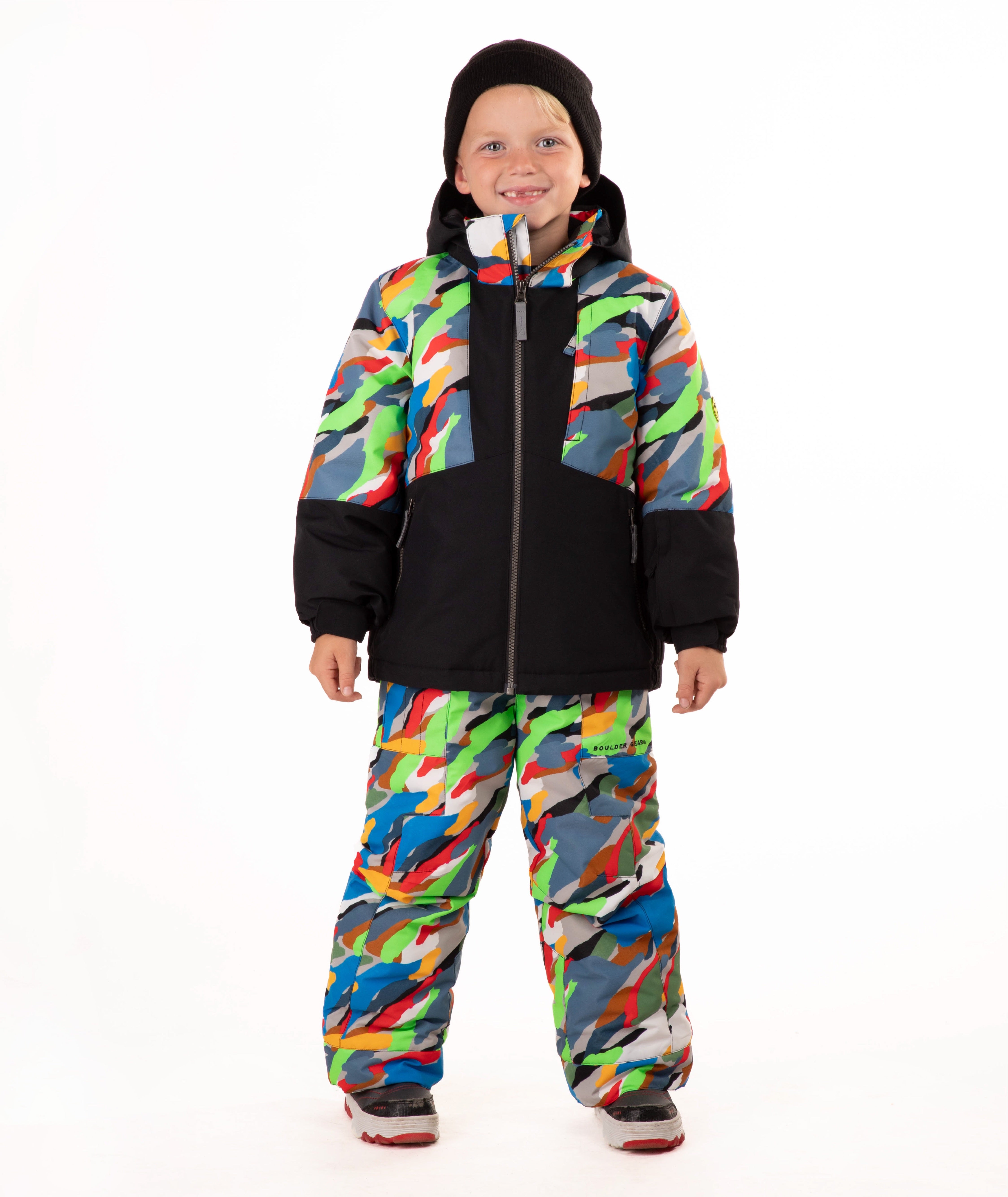 A child wearing a colorful camouflage ski jacket and matching pants with a black beanie, standing against a plain white background.