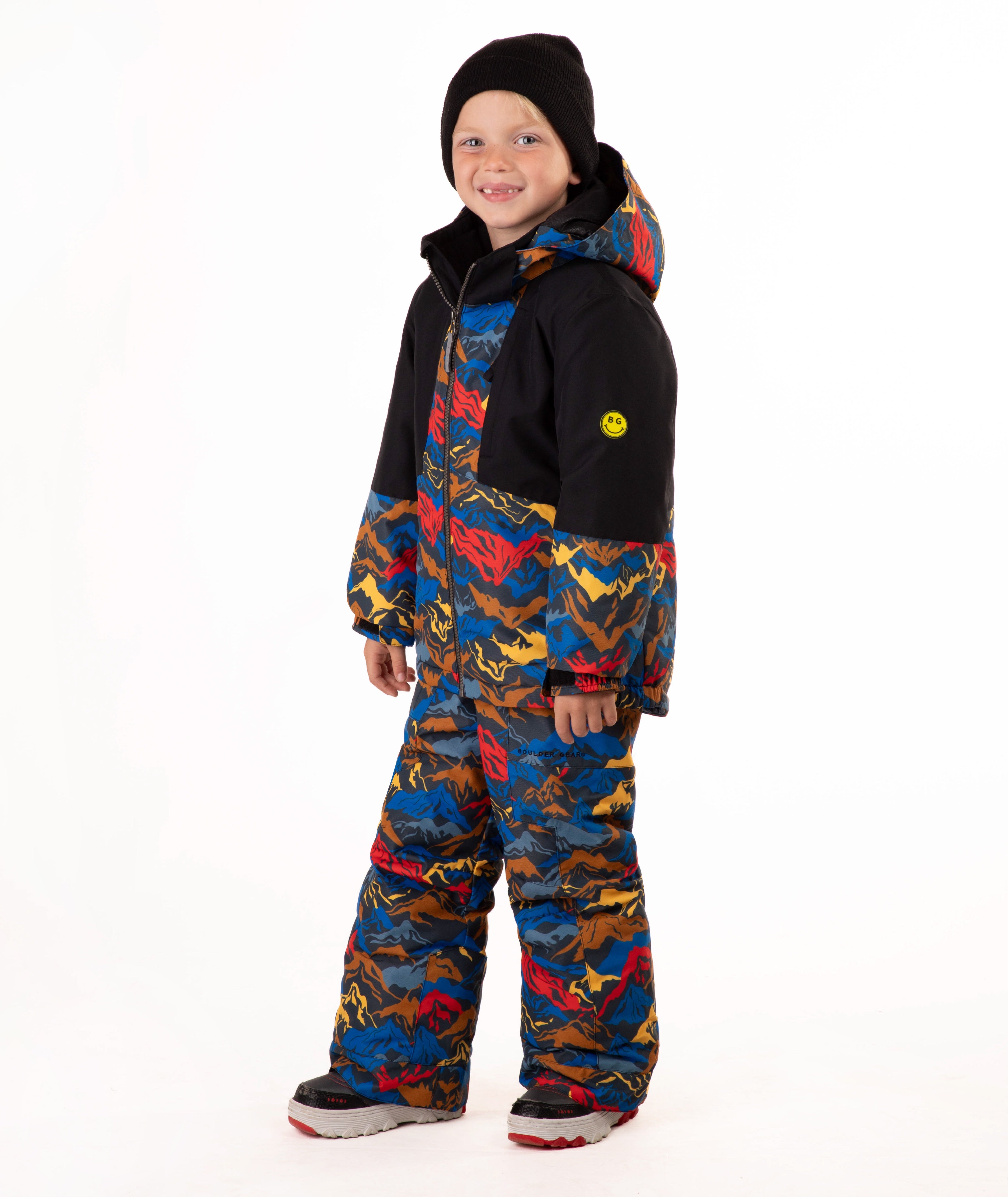 A child wearing a colorful mountain pattern ski jacket and matching pants with a black beanie, standing against a plain white background.