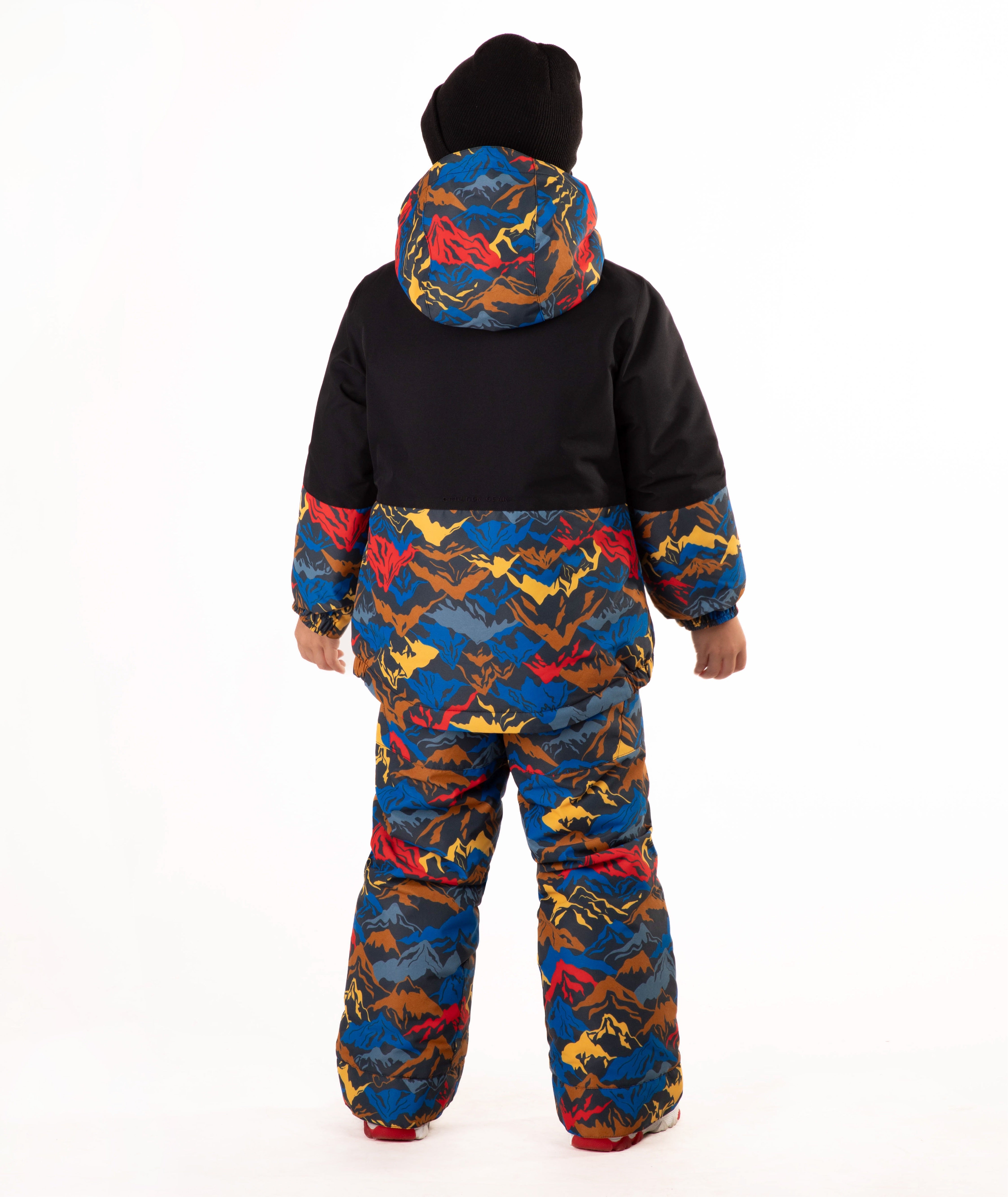 A child wearing a colorful mountain pattern ski jacket and matching pants with a black beanie, standing against a plain white background.