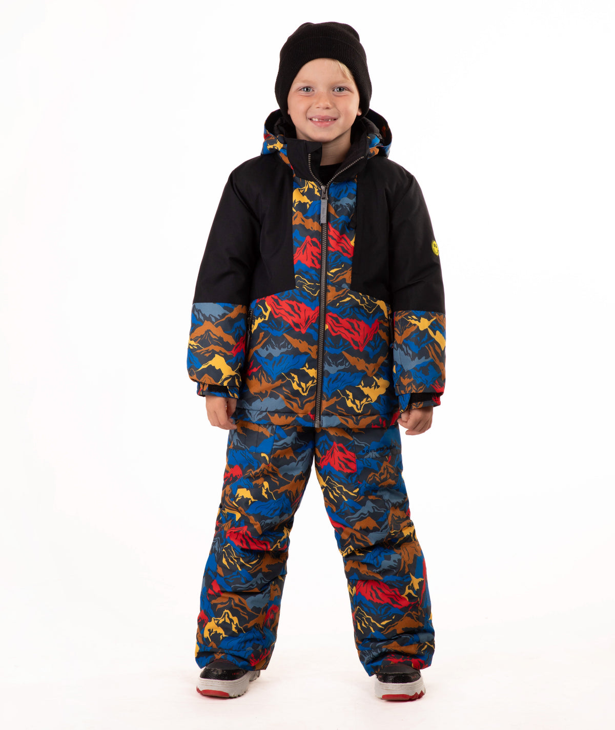 A child wearing a colorful mountain pattern ski jacket and matching pants with a black beanie, standing against a plain white background.