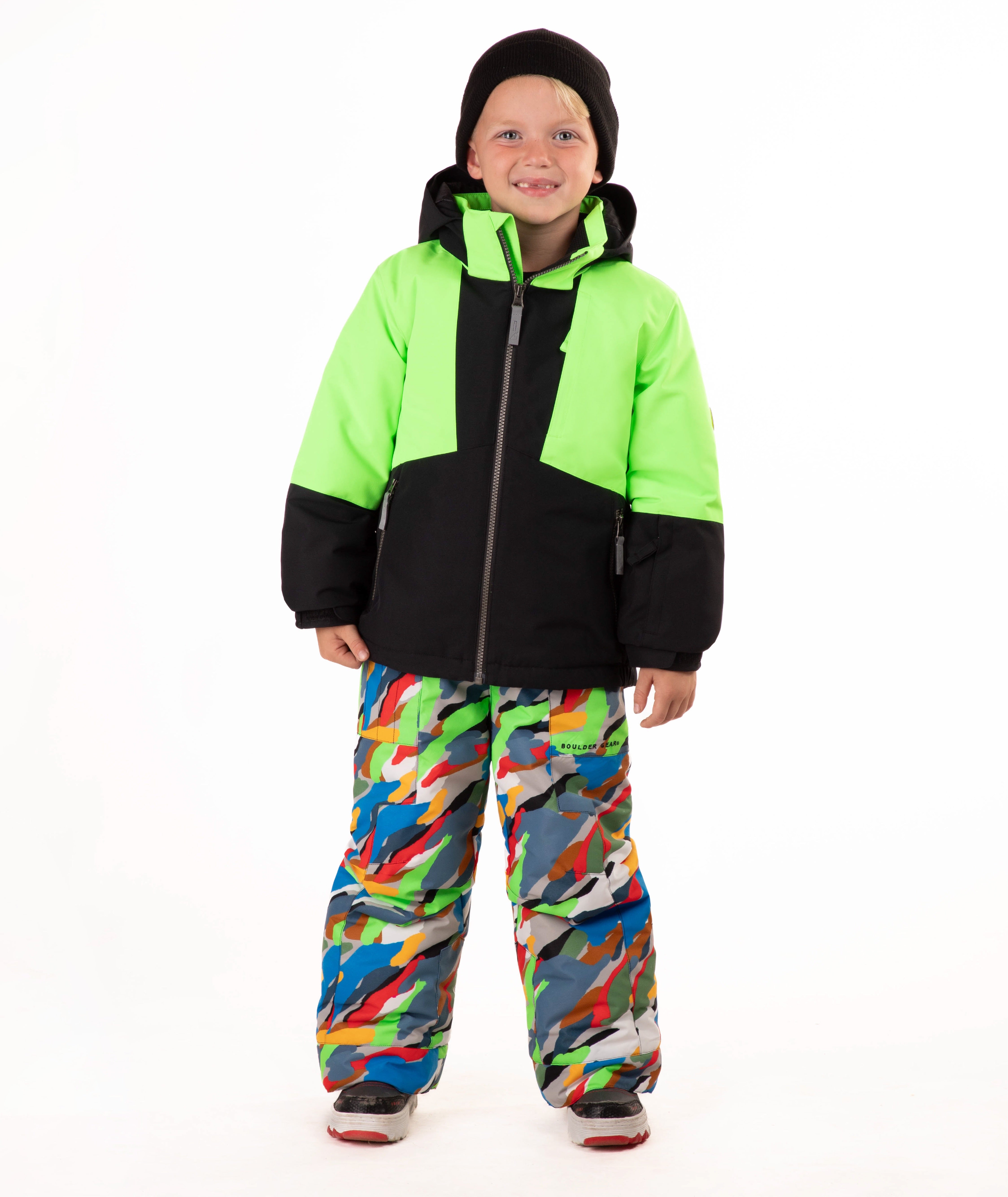 A child wearing a bright neon green and black ski jacket with colorful camouflage snow pants, a black beanie, and ski boots, standing against a plain white background.
