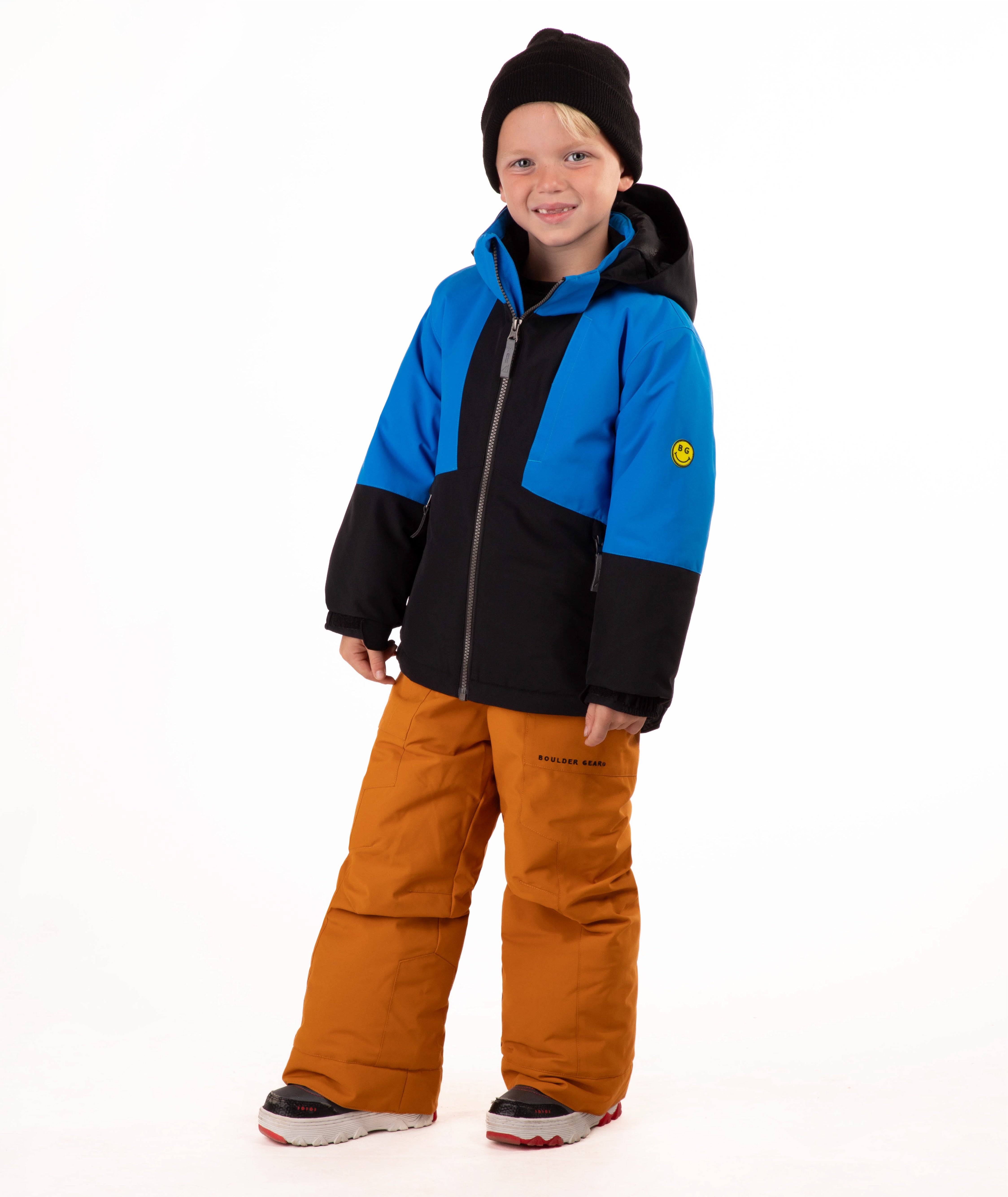 A child standing against a white studio background wearing a color-blocked ski jacket in blue and black, brown snow pants, and a black beanie