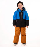 A child standing against a white studio background wearing a color-blocked ski jacket in blue and black, brown snow pants, and a black beanie