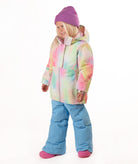 A young girl stands against a white studio background wearing a pastel tie-dye hooded snow jacket, light blue snow pants, and a purple beanie.