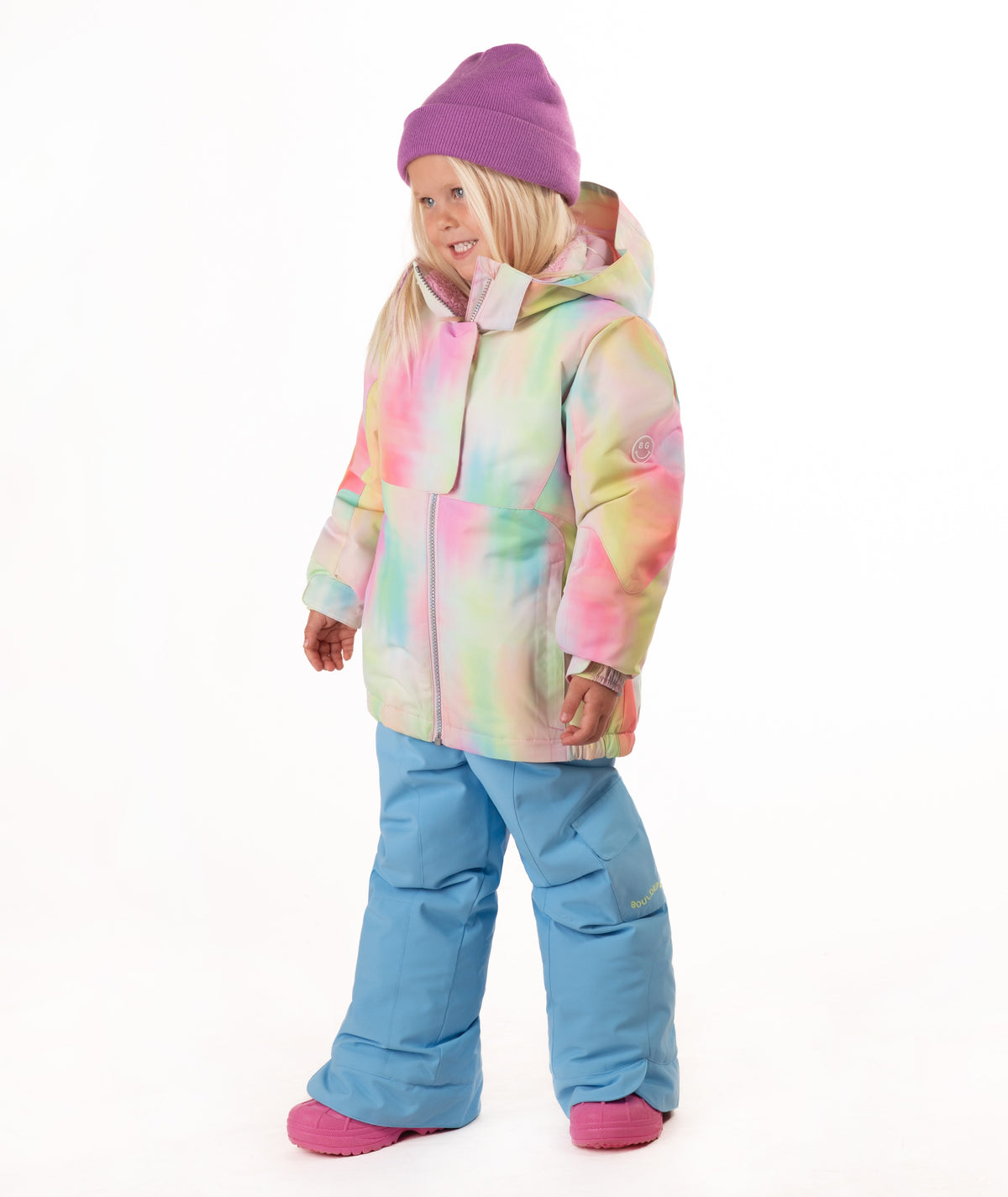 A young girl stands against a white studio background wearing a pastel tie-dye hooded snow jacket, light blue snow pants, and a purple beanie.