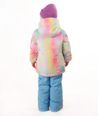 A young girl stands against a white studio background wearing a pastel tie-dye hooded snow jacket, light blue snow pants, and a purple beanie.