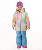 A young girl stands against a white studio background wearing a pastel tie-dye hooded snow jacket, light blue snow pants, and a purple beanie.