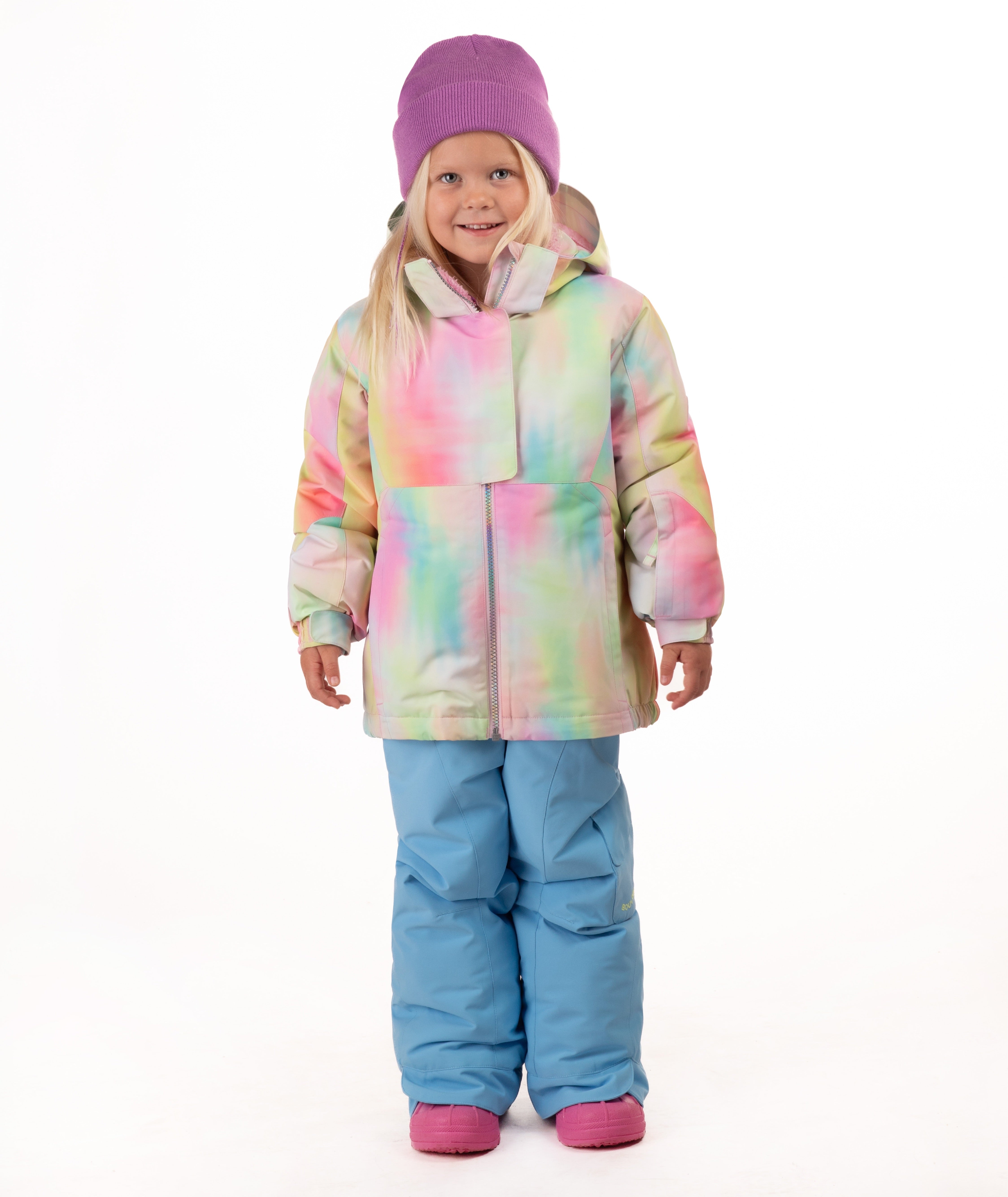 A young girl stands against a white studio background wearing a pastel tie-dye hooded snow jacket, light blue snow pants, and a purple beanie.