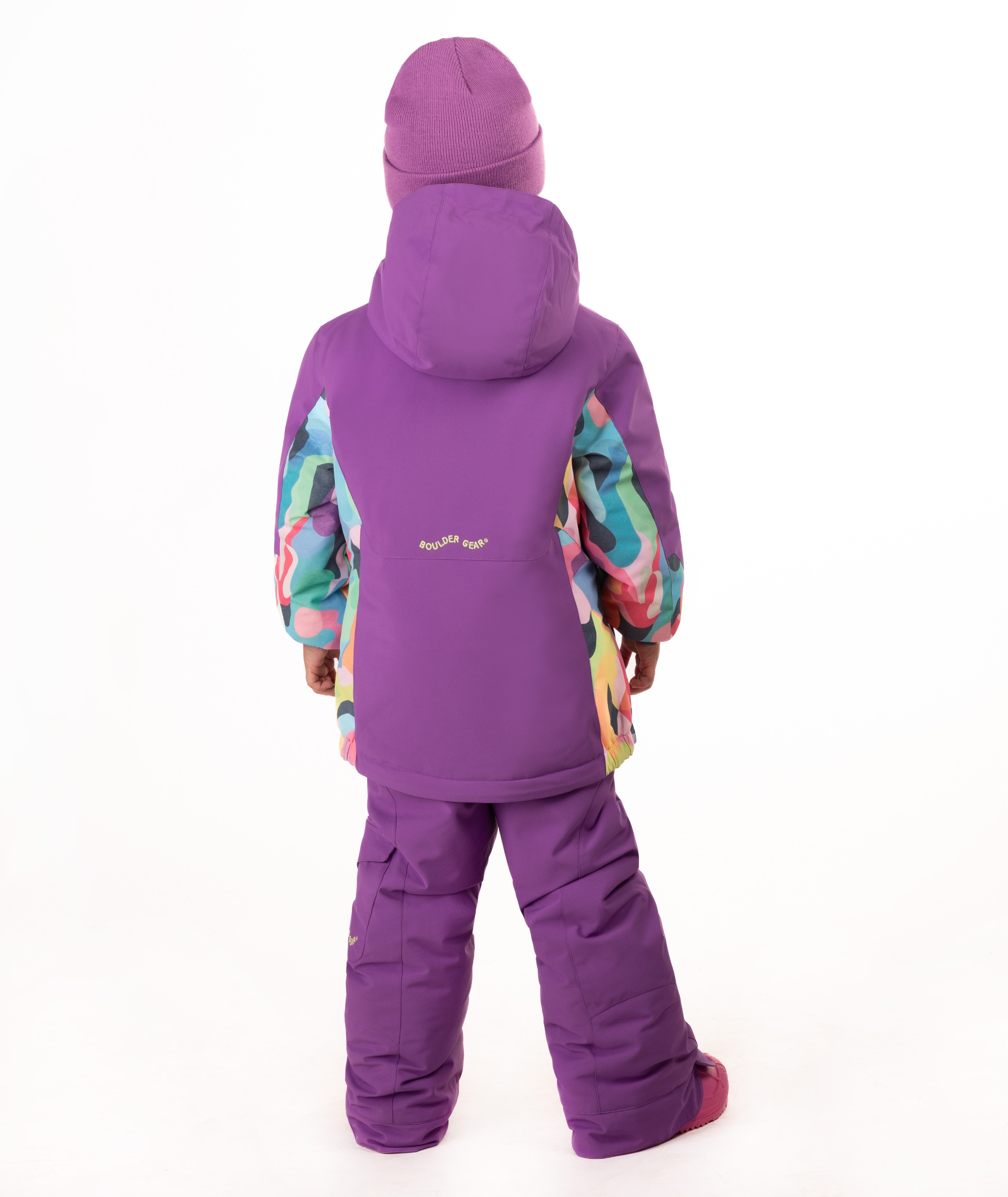 A young girl wearing a purple ski outfit with colorful geometric patterned accents on the sleeves, a matching purple beanie, and purple snow pants, standing against a white background.