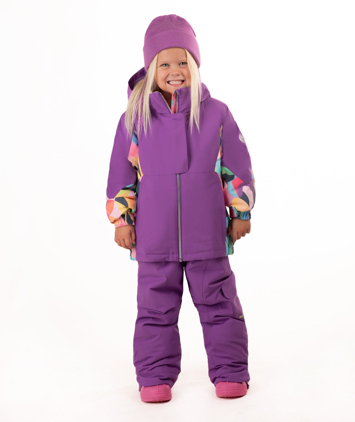 A young girl wearing a purple ski outfit with colorful geometric patterned accents on the sleeves, a matching purple beanie, and purple snow pants, standing against a white background.