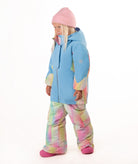 A young child wearing a light blue ski jacket with pastel tie-dye panels, matching tie-dye snow pants, a pink beanie, and pink boots, standing against a plain white background.