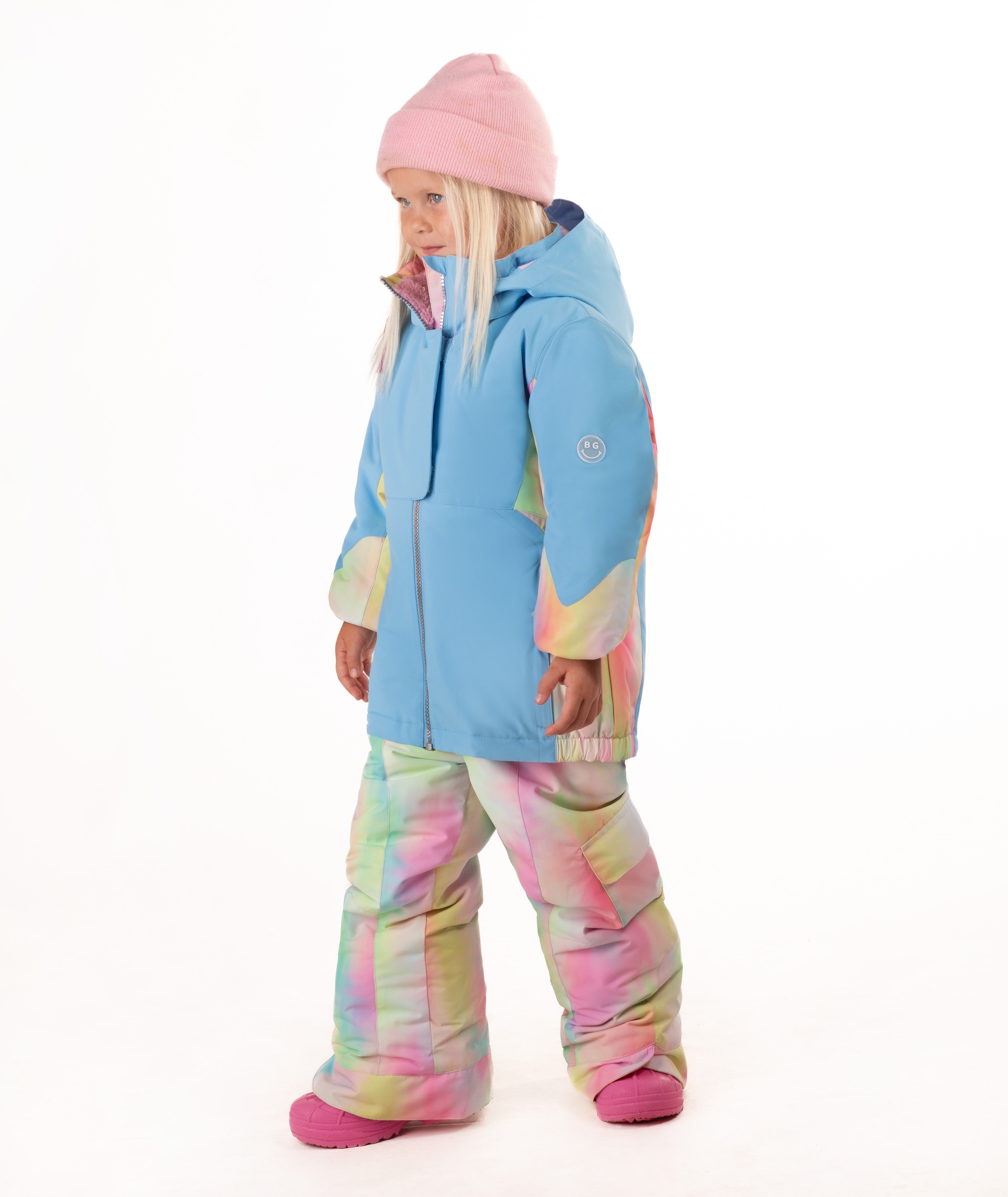A young child wearing a light blue ski jacket with pastel tie-dye panels, matching tie-dye snow pants, a pink beanie, and pink boots, standing against a plain white background.