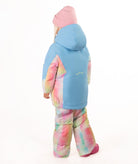 A young child wearing a light blue ski jacket with pastel tie-dye panels, matching tie-dye snow pants, a pink beanie, and pink boots, standing against a plain white background.