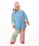A young child wearing a light blue ski jacket with pastel tie-dye panels, matching tie-dye snow pants, a pink beanie, and pink boots, standing against a plain white background.