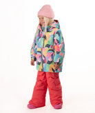 A young child wearing a colorful abstract-patterned ski jacket, bright pink snow pants, and a pink beanie, standing against a plain white studio background.