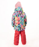A young child wearing a colorful abstract-patterned ski jacket, bright pink snow pants, and a pink beanie, standing against a plain white studio background.