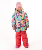A young child wearing a colorful abstract-patterned ski jacket, bright pink snow pants, and a pink beanie, standing against a plain white studio background.