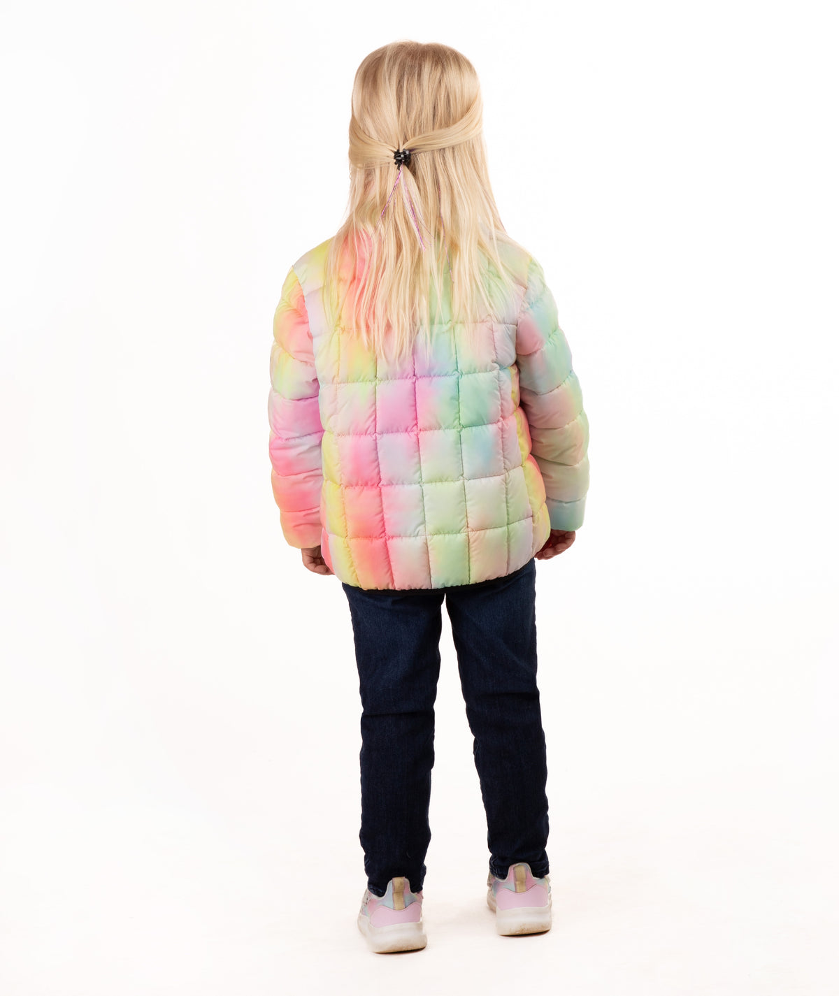 A young blonde girl wearing a pastel rainbow puffer jacket, dark blue jeans, and gray sneakers, standing against a solid white background.