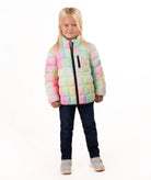 A young blonde girl wearing a pastel rainbow puffer jacket, dark blue jeans, and gray sneakers, standing against a solid white background.