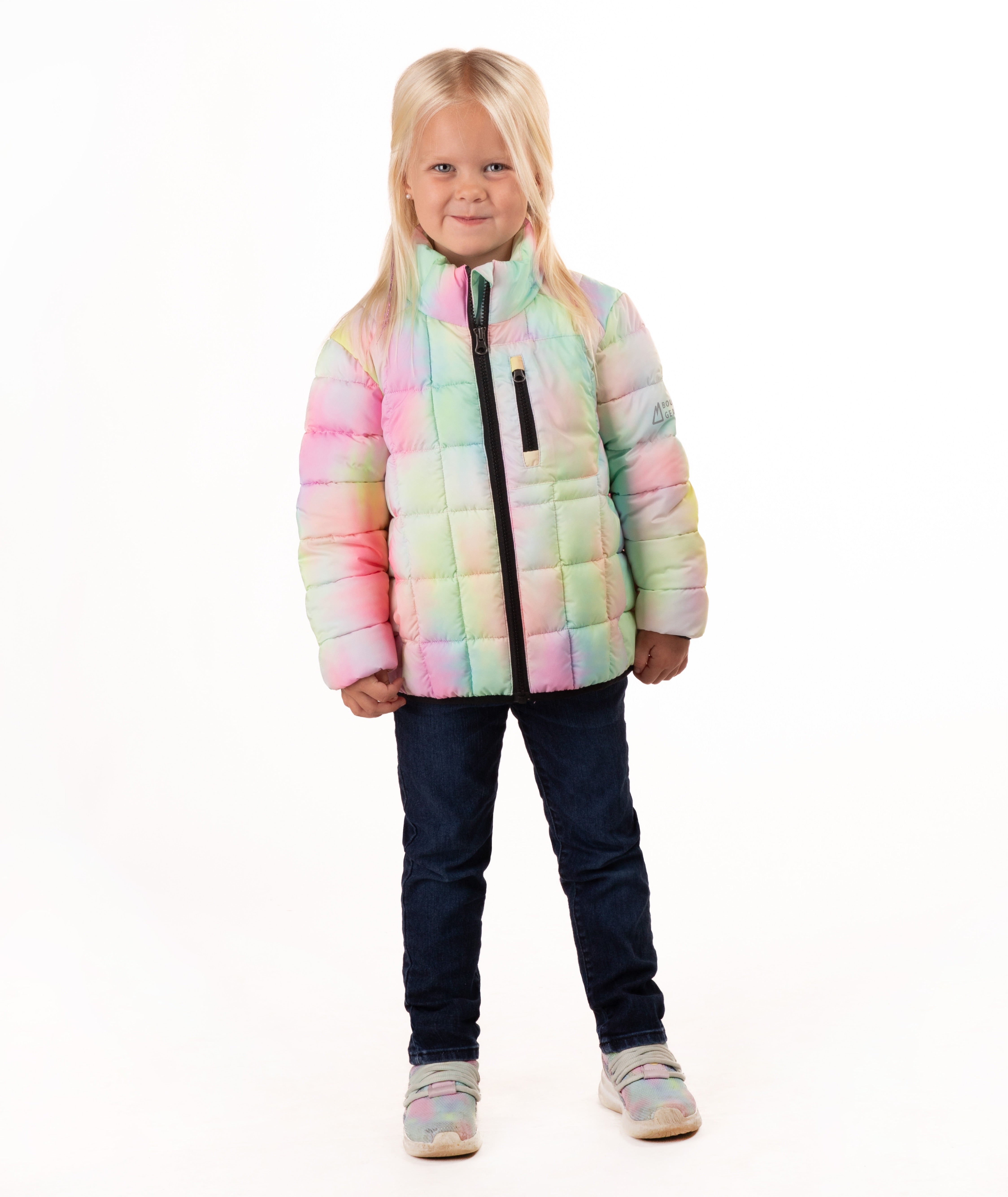 A young blonde girl wearing a pastel rainbow puffer jacket, dark blue jeans, and gray sneakers, standing against a solid white background.