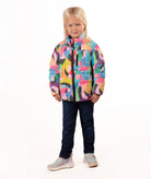 A young girl with blonde hair stands against a white studio background wearing a colorful abstract print puffer jacket, dark blue jeans, and light gray sneakers.