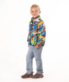 A young boy stands against a white studio background wearing a colorful puffer jacket with a multi color camo print, blue jeans, and brown shoes.
